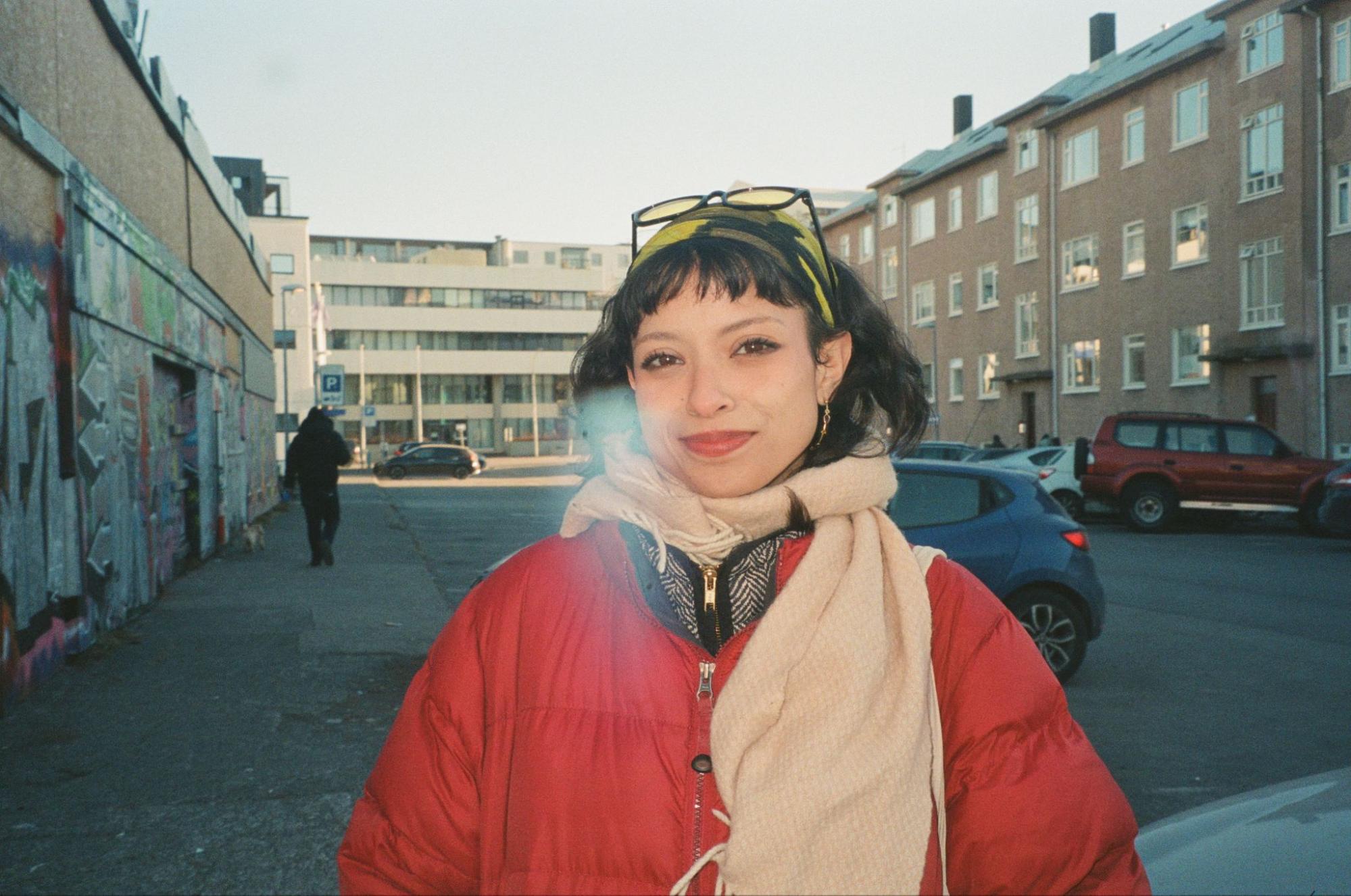 A film photo of Audrey taken outside in Reykjavik, Iceland. She is wearing a red puffer coat, cream wool scarf, green headband, and sunglasses on top of her head. There are buildings and cars in the background.